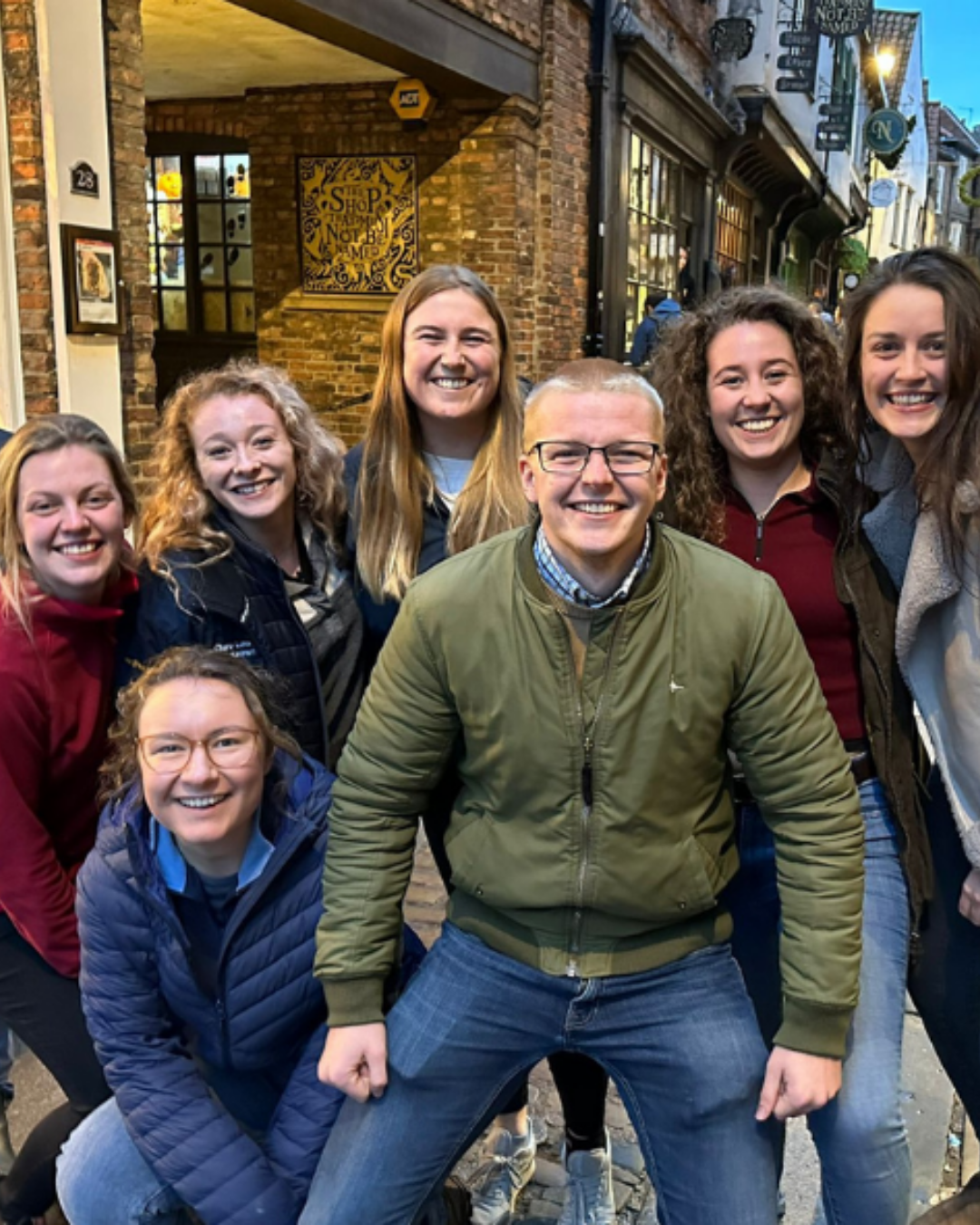 A group of seven smiling friends poses for a photo on a cobblestone street, with charming brick buildings in the background. They are wearing casual clothing and appear happy and vibrant.