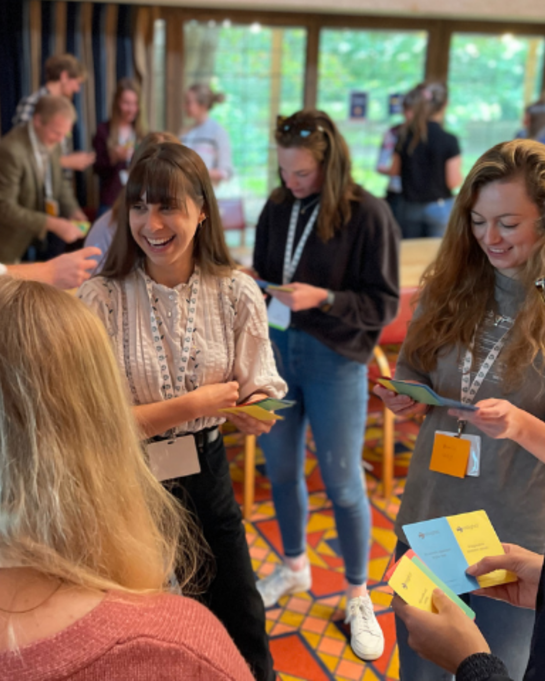 Participants engage in a group activity, smiling and holding colorful cards. A few people in the background are also interacting, while the setting has a vibrant, patterned floor.